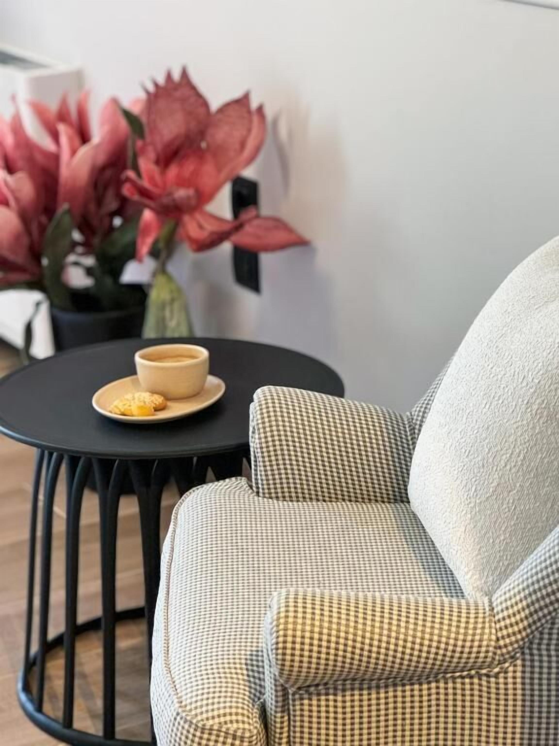 Un fauteuil à carreaux avec un coussin, une petite table ronde avec une tasse et une soucoupe, et un pot de fleurs orné de grandes fleurs roses.
