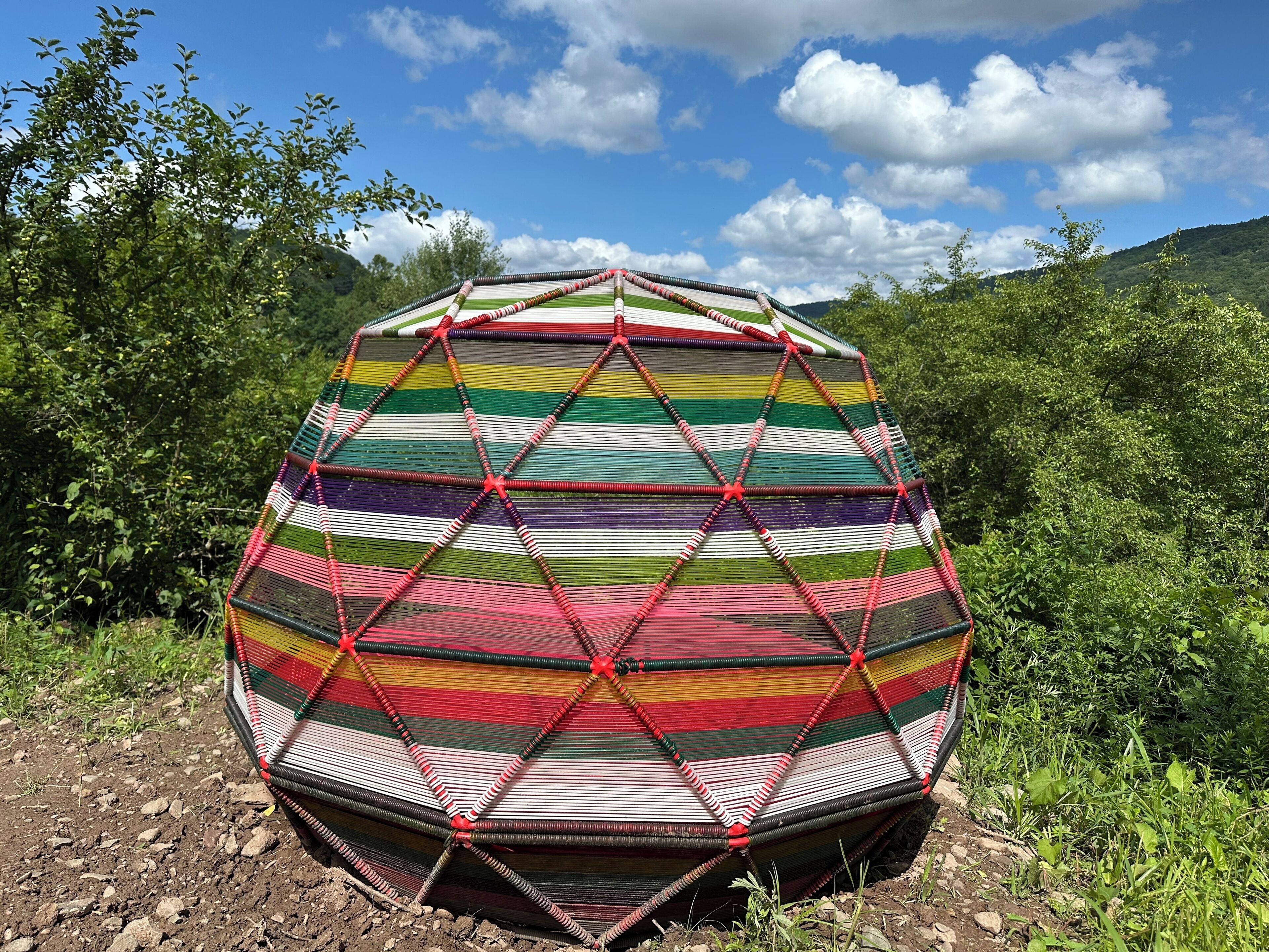 Una estructura de cúpula geodésica con un patrón colorido a rayas, rodeada de follaje y un cielo despejado.