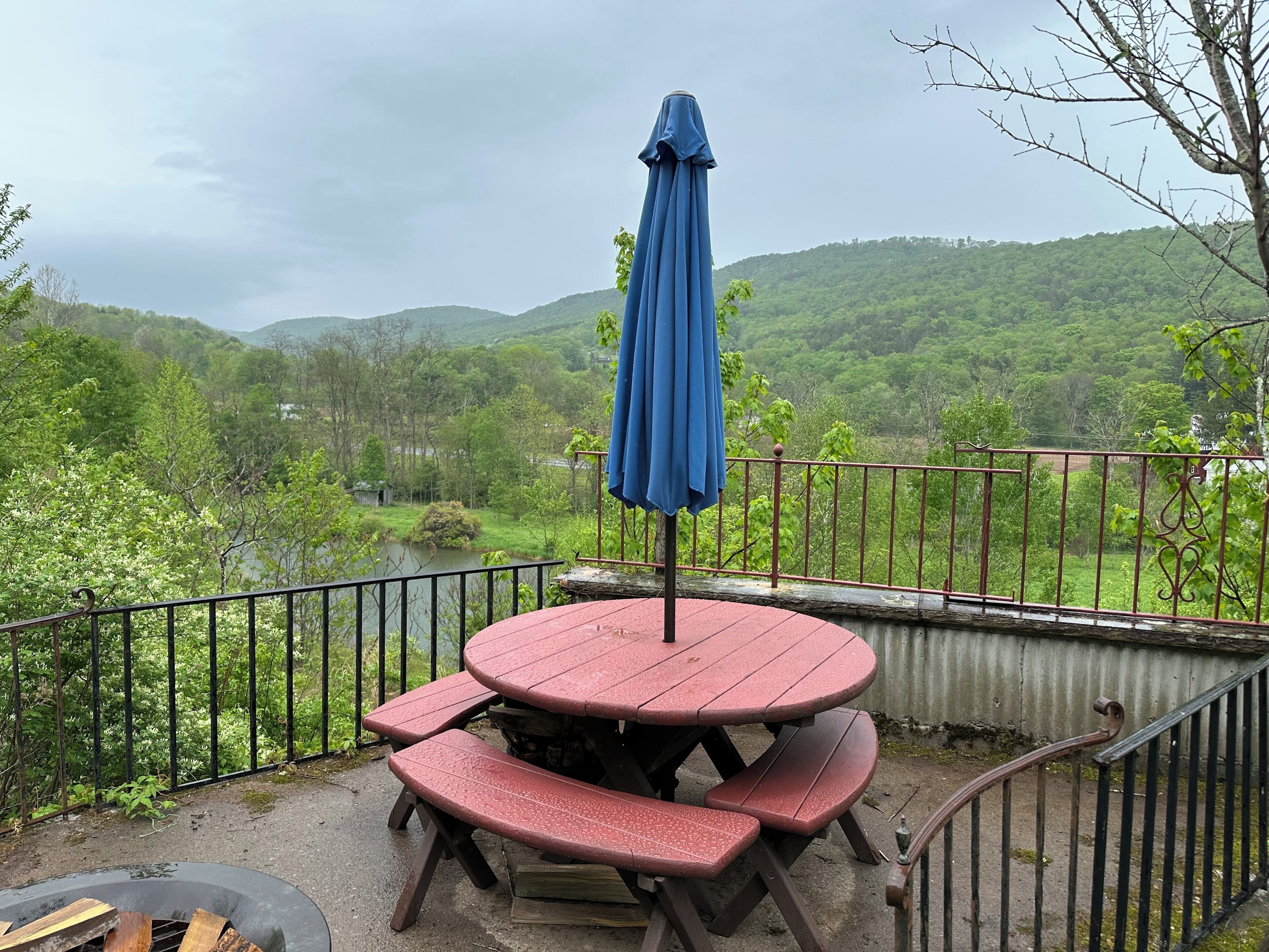 A red picnic table with benches and a blue umbrella on a balcony overlooking a river and green hills.