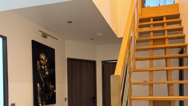 Een modern interieur met een houten trap, een kunstwerk in een lijst aan de muur, en een deur aan het einde van de gang.