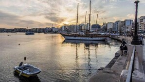 A waterfront scene with a large sailing ship docked, a smaller boat, and a cityscape in the background. - Beautiful 3BR home Apartment in Gzira w/WIFI & AC (Il-Gżira)