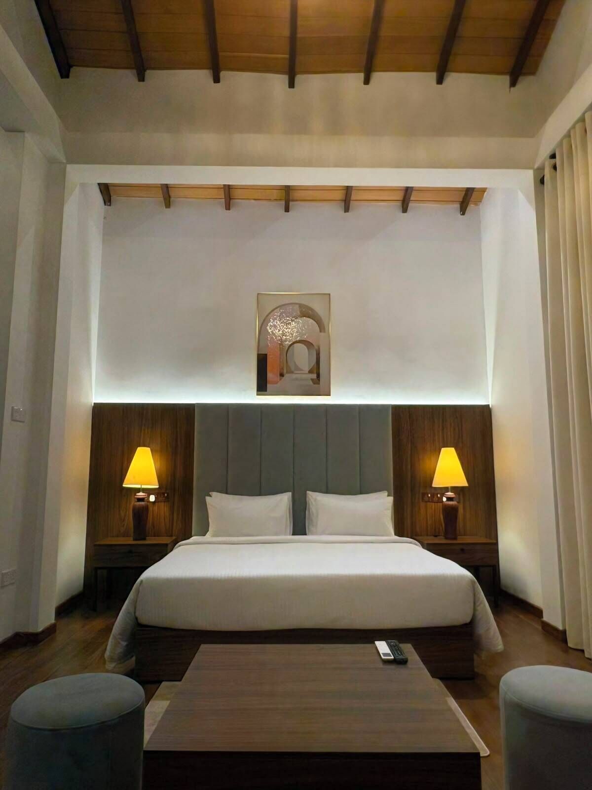 A hotel room with a large bed, two bedside tables with lamps, a wooden headboard, and a ceiling with wooden beams.