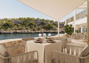 Área de refeições ao ar livre com uma mesa posta para dois, com vista para a costa rochosa e árvores.