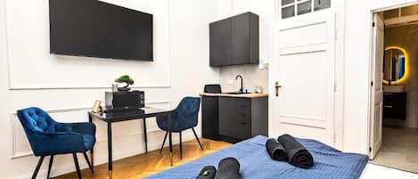 Quarto compacto com cama, escrivaninha e cadeira, televisão e pequena cozinha.