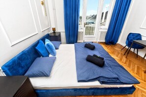 Ein Schlafzimmer mit einem Bett, blauer Bettwäsche, einem Nachttisch, einem Sessel und Blick ins Freie.