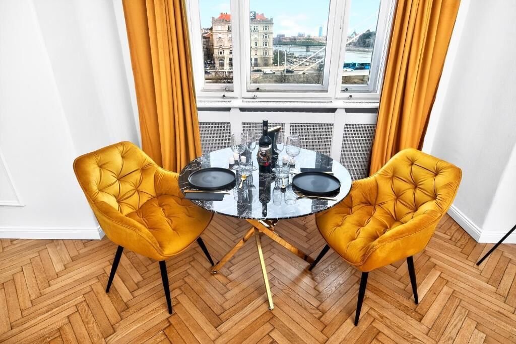 A dining area with a round table and two yellow chairs, a window with curtains, and a view of buildings.