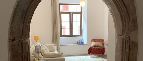 An interior space with a stone archway leading to a room with a sofa, a small table, and a window with curtains.