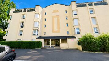 Un hôtel à plusieurs étages, avec une entrée principale, un parking et une voiture garée au premier plan.
