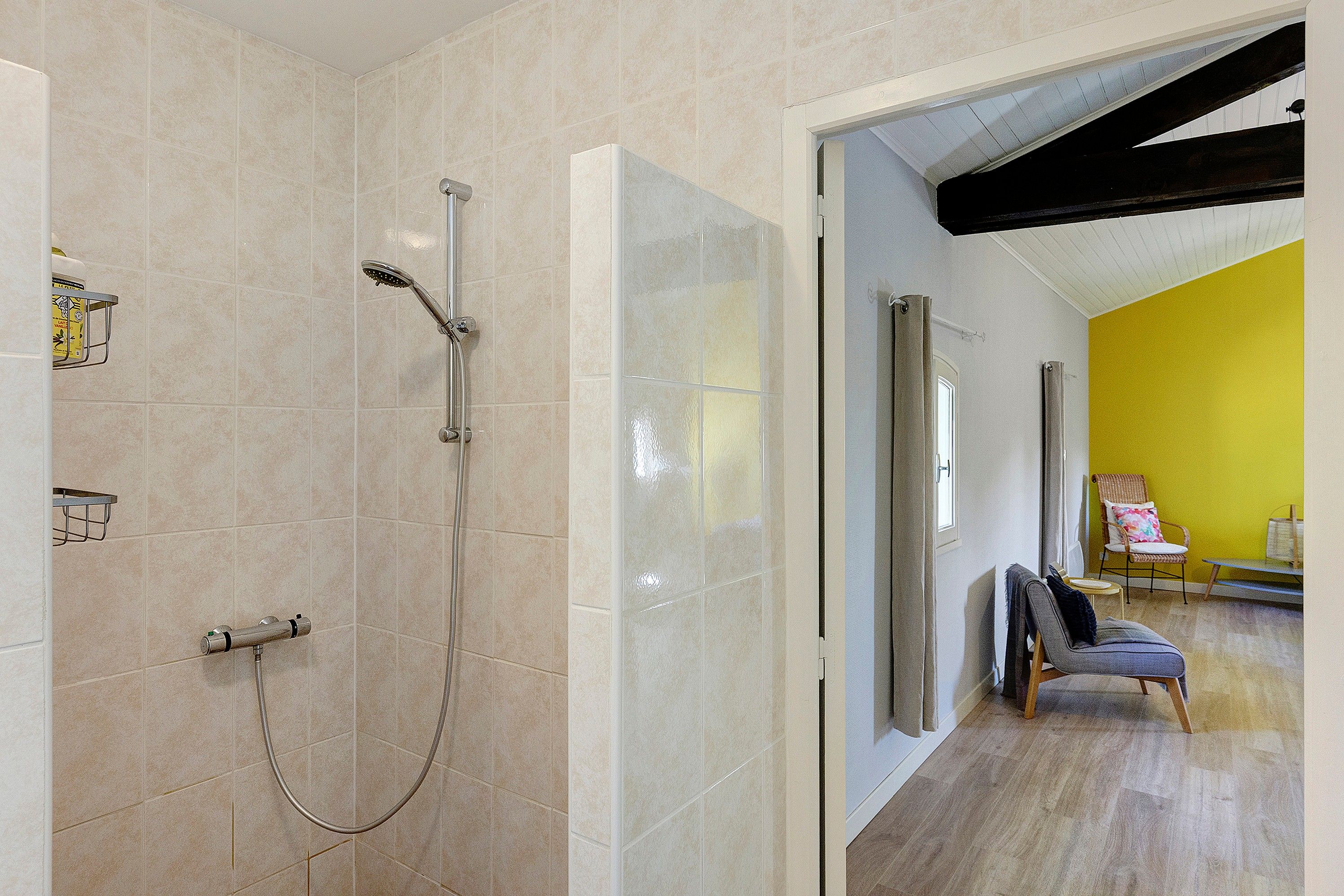 Un baño moderno con ducha, inodoro y lavabo. Se ve una puerta que da a una zona de estar con una pared amarilla y una silla.