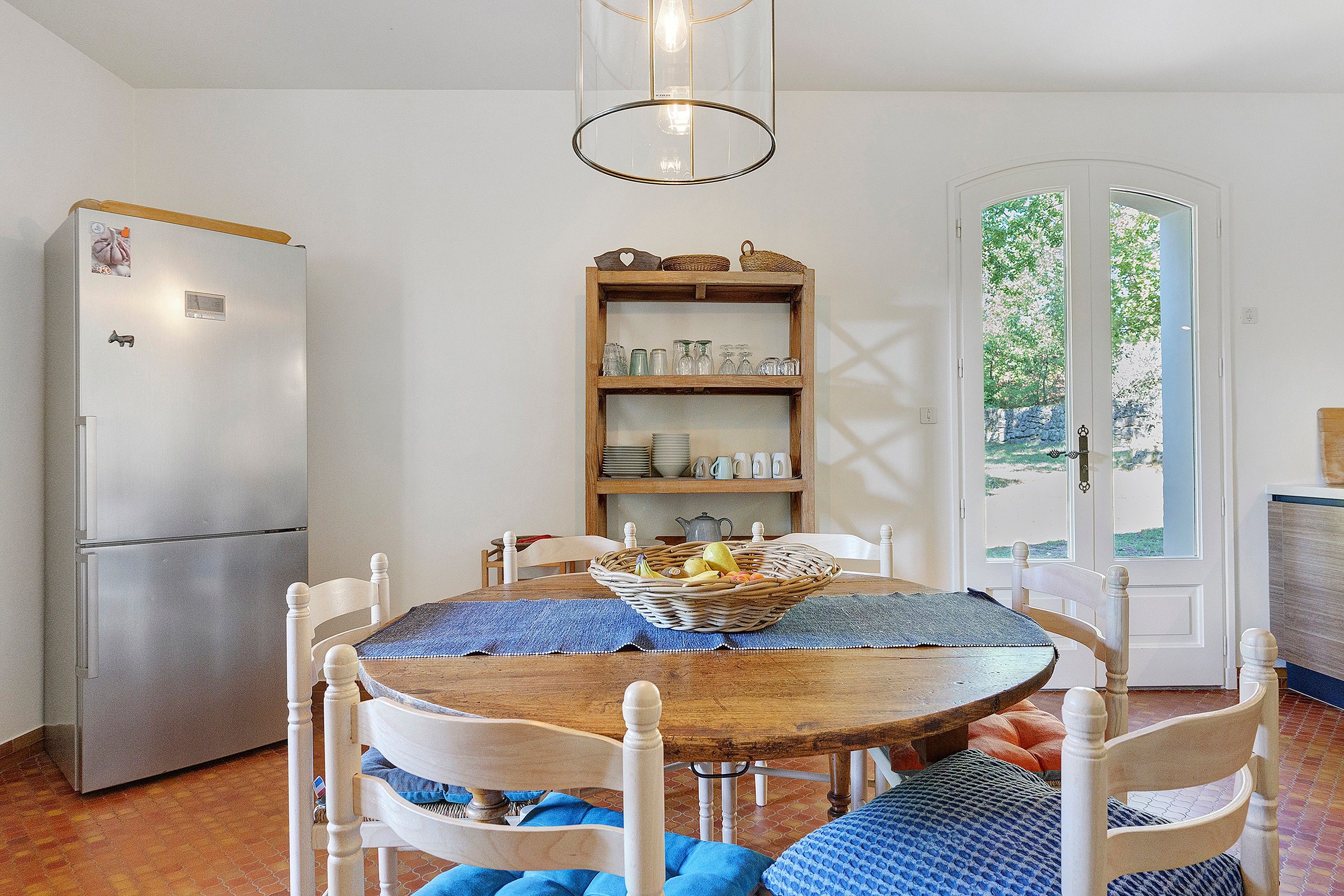 Uma sala de jantar com mesa de madeira, cadeiras, geladeira, prateleira com copos e uma porta com vista.