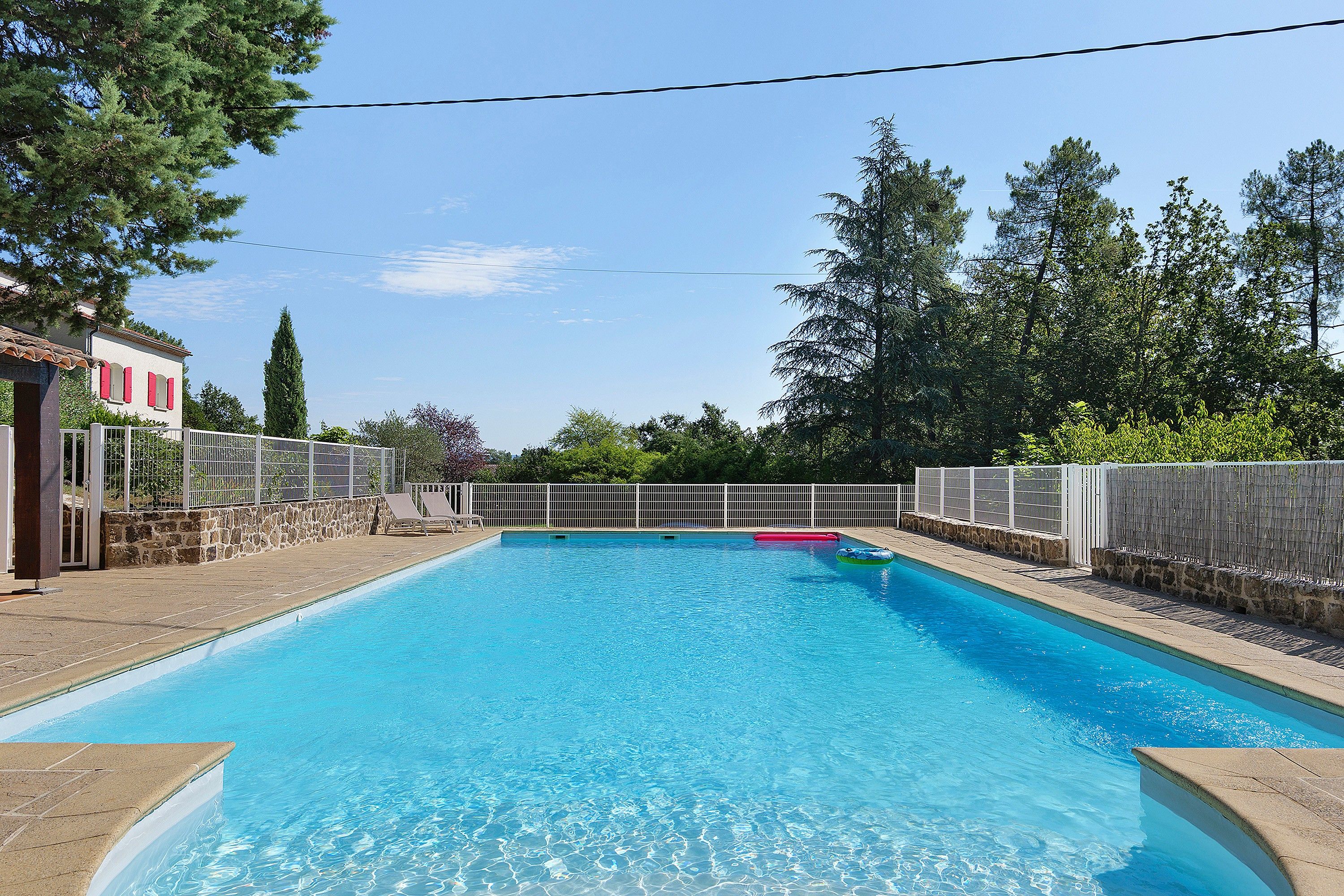 Una piscina rectangular con agua azul cristalina, rodeada por una solana de piedra y una valla blanca.