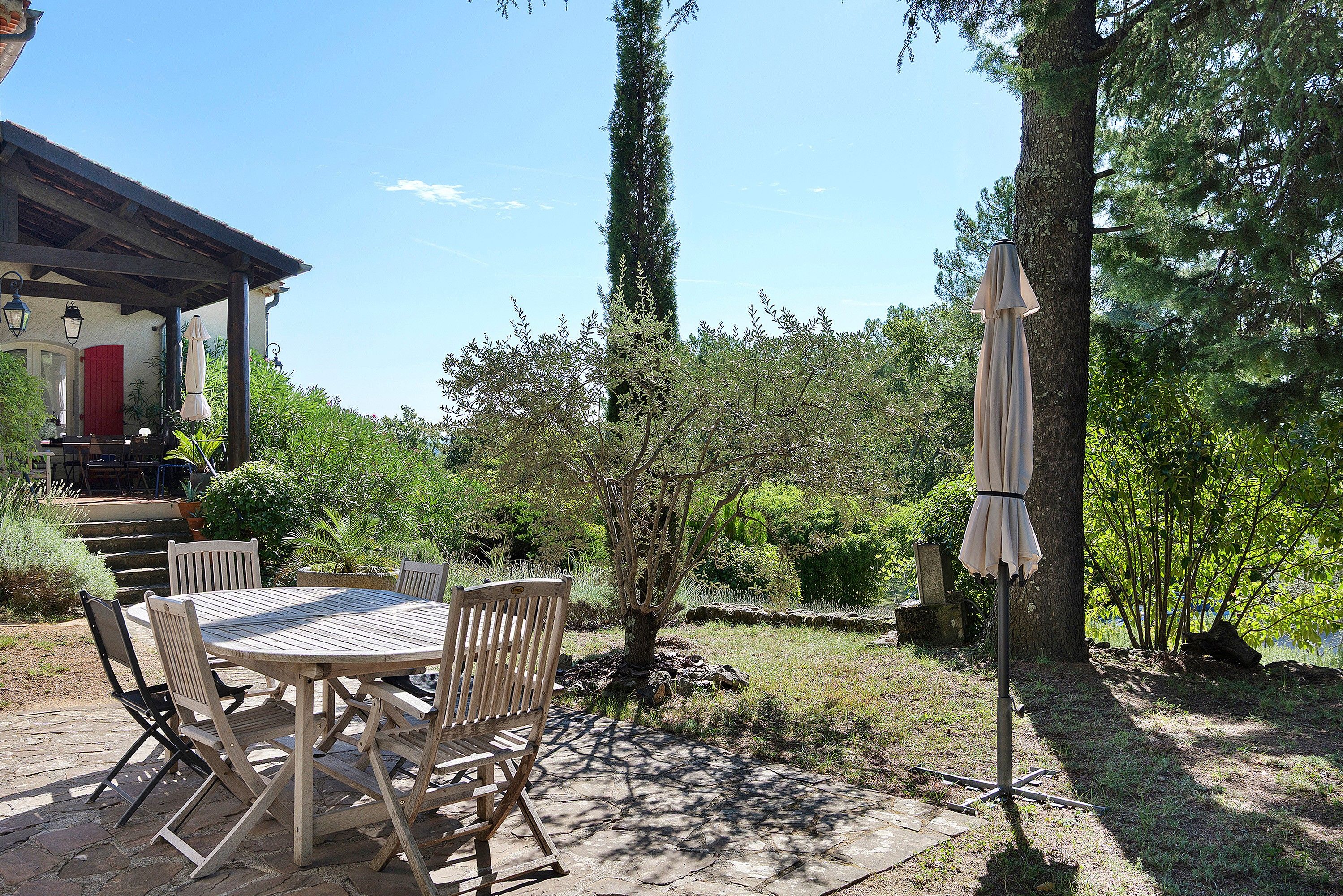 Eine Terrasse im Freien mit einem runden Tisch und Stühlen, umgeben von Grünpflanzen und Bäumen.