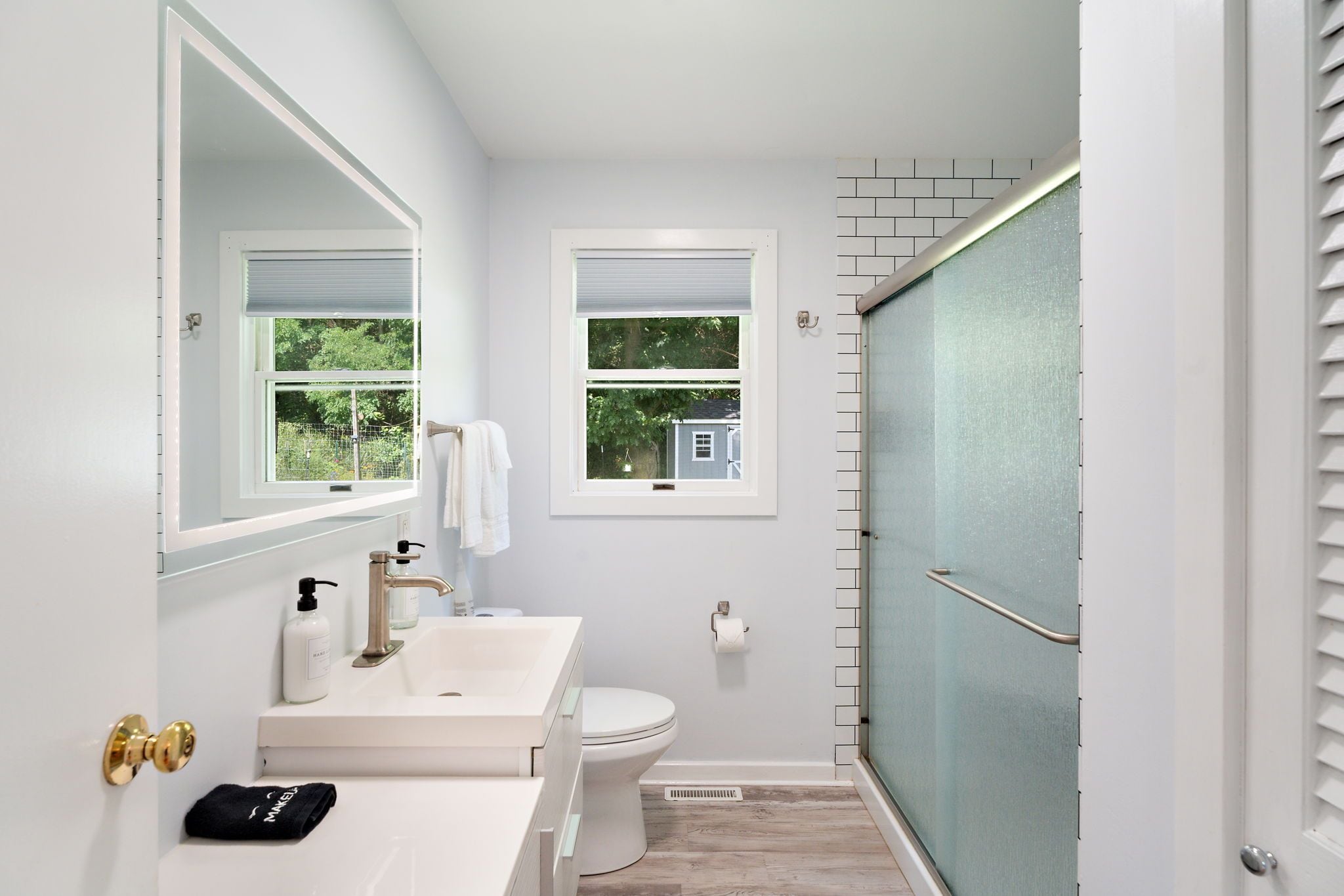 Un baño con lavamanos, inodoro y ducha blancos. Hay dos ventanas con vistas a zonas verdes.
