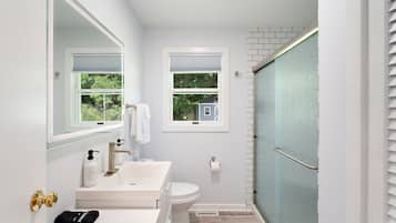 Un baño con lavamanos, inodoro y ducha blancos. Hay dos ventanas con vistas a zonas verdes.