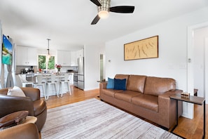 A modern living room with a brown leather sofa, a dining area with white chairs, a kitchen with white cabinets, and a ceiling fan.