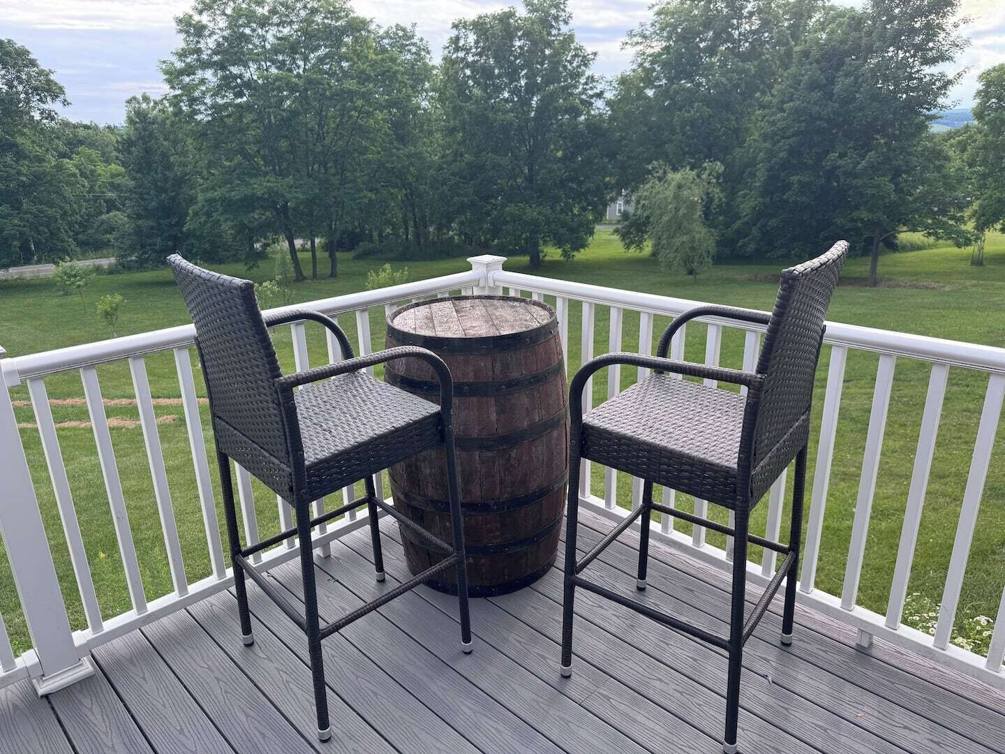 A deck with two wicker chairs and a wooden barrel, surrounded by a white railing, overlooking a grassy area.