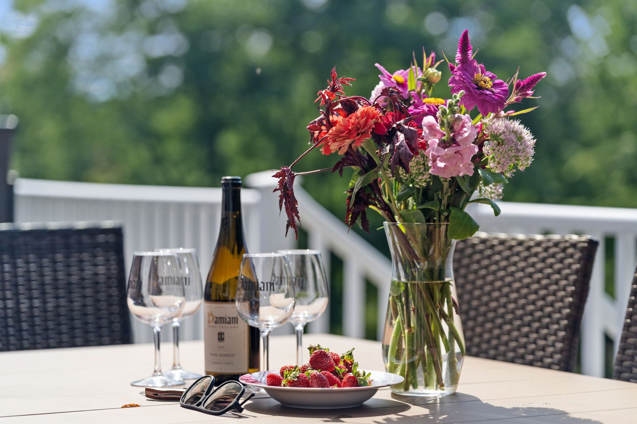 A table with a bottle of wine, two wine glasses, a vase with flowers, a bowl of strawberries, and a pair of sunglasses.
