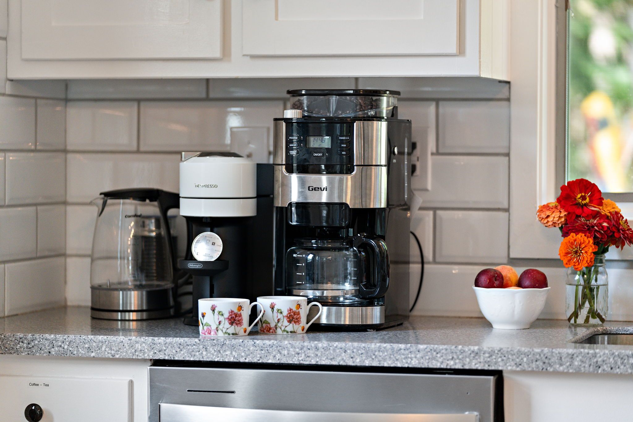 A kitchen with a coffee maker, a glass carafe, two floral mugs, a bowl of fruit, and a vase with flowers.