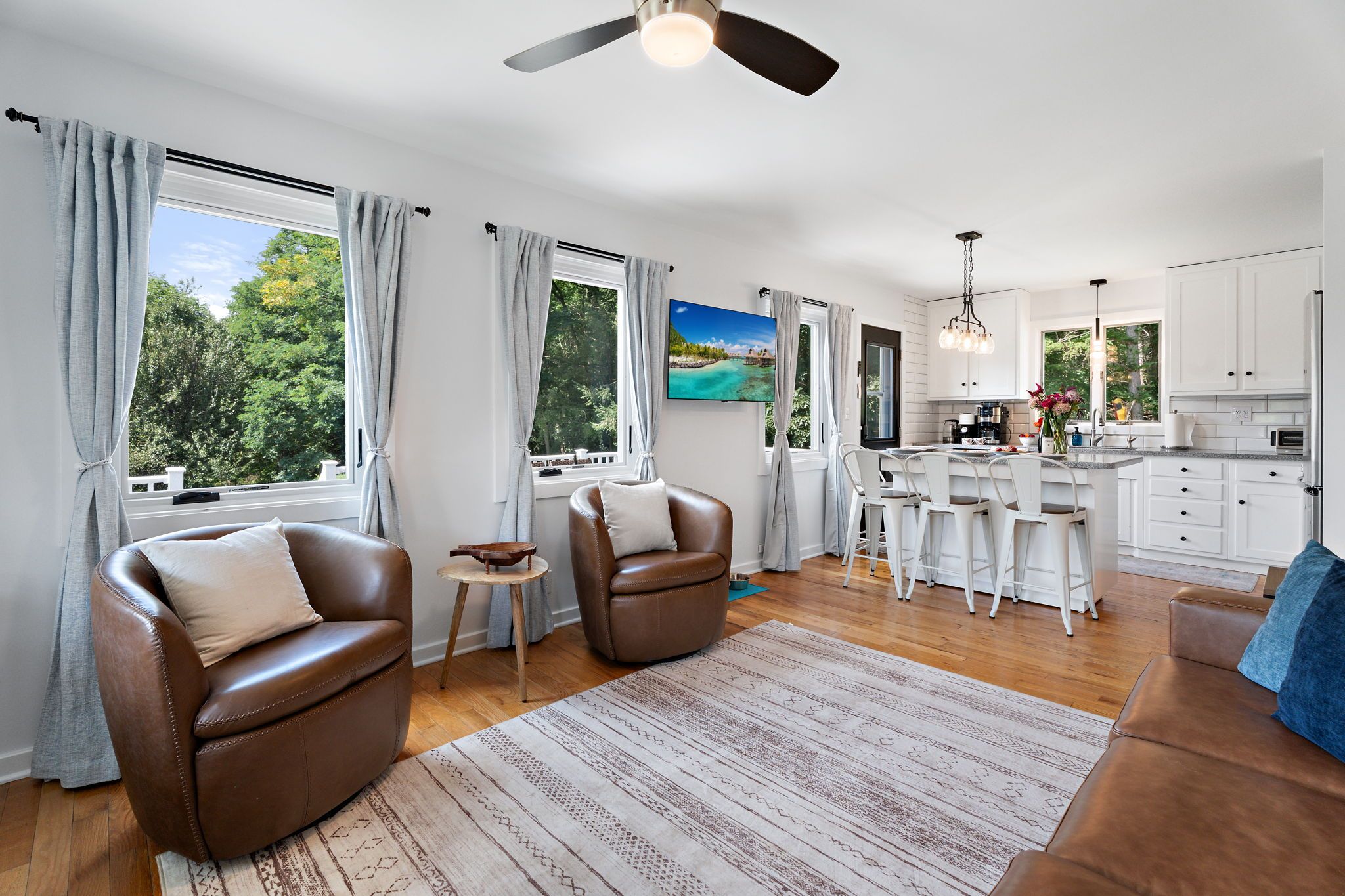 A spacious living area with a large window, a ceiling fan, and a kitchen with white cabinets and a dining area.