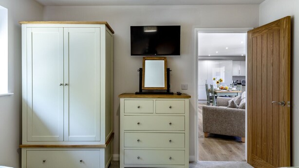 A bedroom with a wardrobe, a dresser, a television, and an open door leading to a living area. - The Hay Barn at Rusty Lane Farm (Seend)