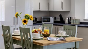A modern kitchen with white cabinets, a wooden dining table set with plates, glasses, and a bowl of fruit. - The Hay Barn at Rusty Lane Farm (Seend)