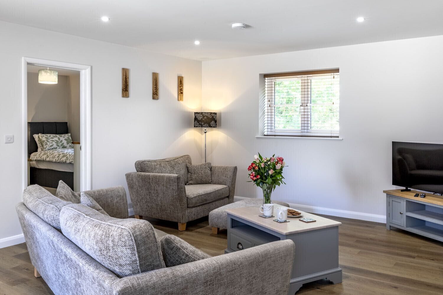 A modern living room with a sofa set, a coffee table with a vase of flowers, a flat-screen TV, and a window with blinds.