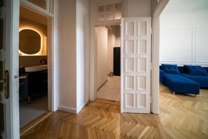 A room with a wooden herringbone floor, a white door, a blue sofa, and a bathroom with a round mirror and sink. - Where the Danube Meets Downtown: Exclusive Scenic Suite – 536 (Budapest)
