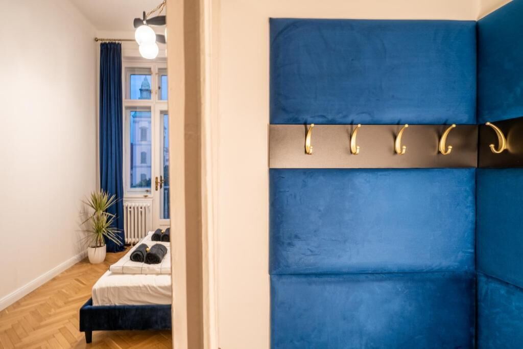 A modern bedroom with a blue upholstered headboard, a bedside table with a lamp, and a view of a building through a window.