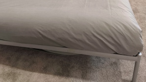 Una cama con colchón gris y dos almohadas, una mesita redonda con lámpara, y piso alfombrado.
