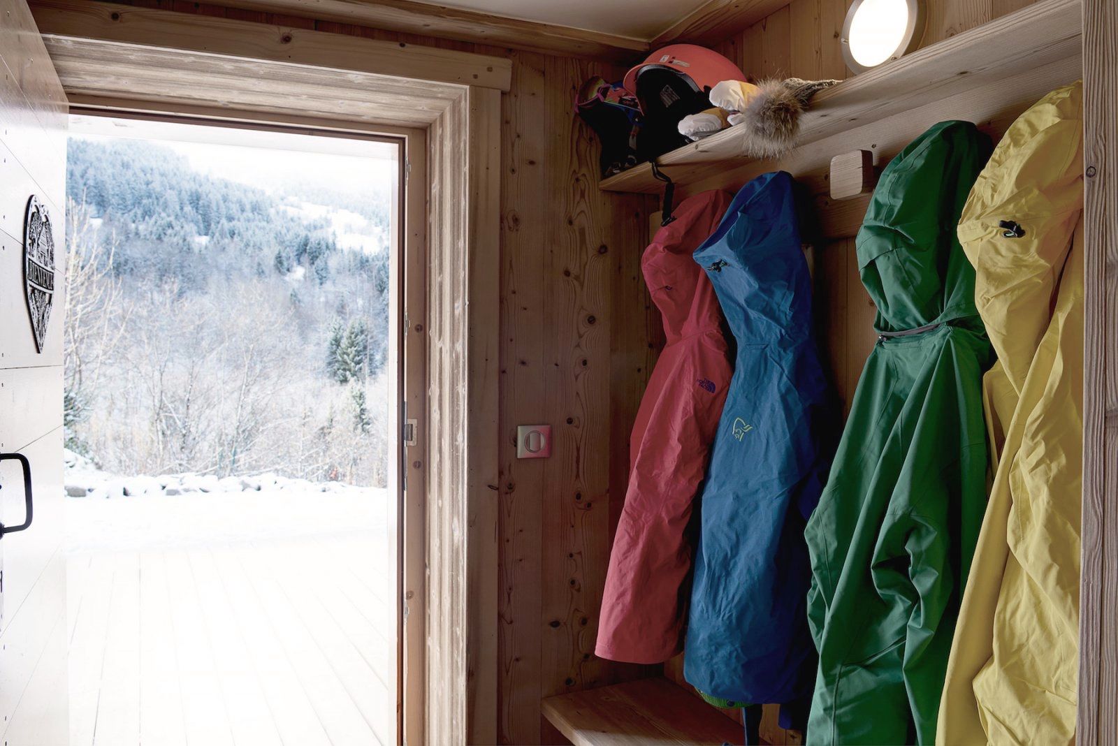 A wooden cabin interior with a door leading to a snowy landscape, a shelf with colorful jackets, and a small shelf with a red bag and a fur item.