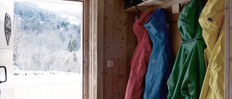 En stuga med träpanel på insidan, med en dörr som leder ut till ett snötäckt landskap. Inuti finns en hylla med färgglada jackor och en mindre hylla med en röd väska och ett föremål av päls.