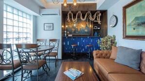 A dining area with a round table and chairs, a bar area with stools, a sofa, and a large window with a view. - Captain Stam Suites Athens - FamilyApartment (Athina)