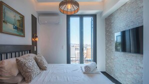 A hotel room with a bed, a painting, a television, and a view of the city through large windows. - Captain Stam Suites Athens - FamilyApartment (Athina)