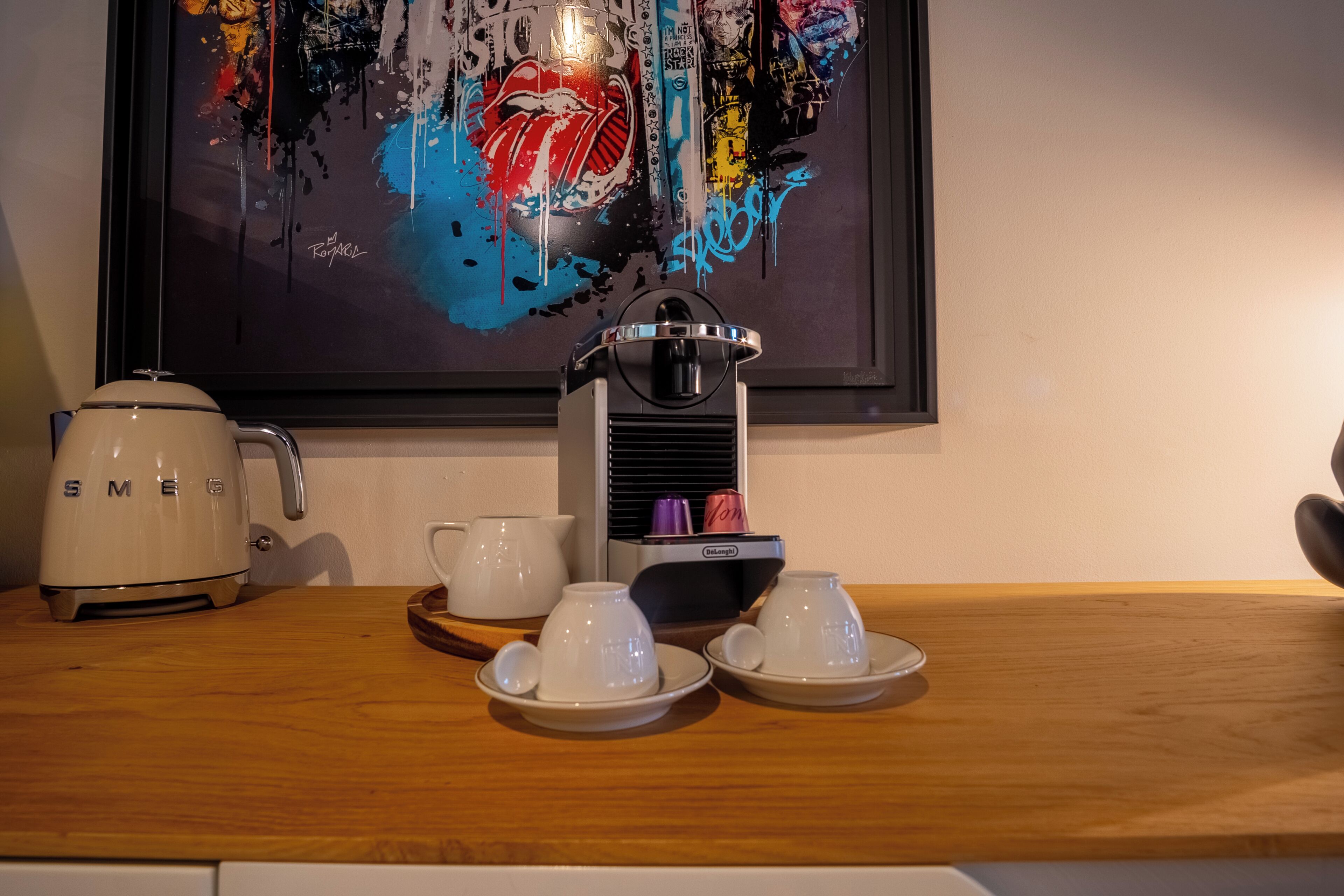 Une machine à café, deux tasses et leurs soucoupes sont posées sur une table en bois, et un tableau encadré est accroché au mur au-dessus.