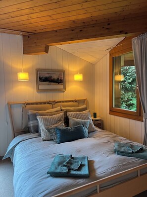 A bedroom with a wooden ceiling, a bed with multiple pillows, a nightstand, a framed picture, and a window with curtains. - Möösli Vue Appartement/Fewo Appartement/Fewo by Interhome (Beatenberg)