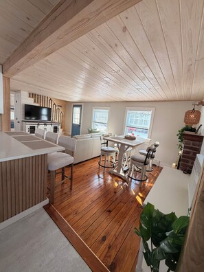A spacious living area with a wooden ceiling, a kitchen with a flat-screen TV, a dining table with chairs, and a large window. - 3-bedroom cottage in Swifts Beach, relaxing coastal living, outdoor shower, AC. (Wareham)