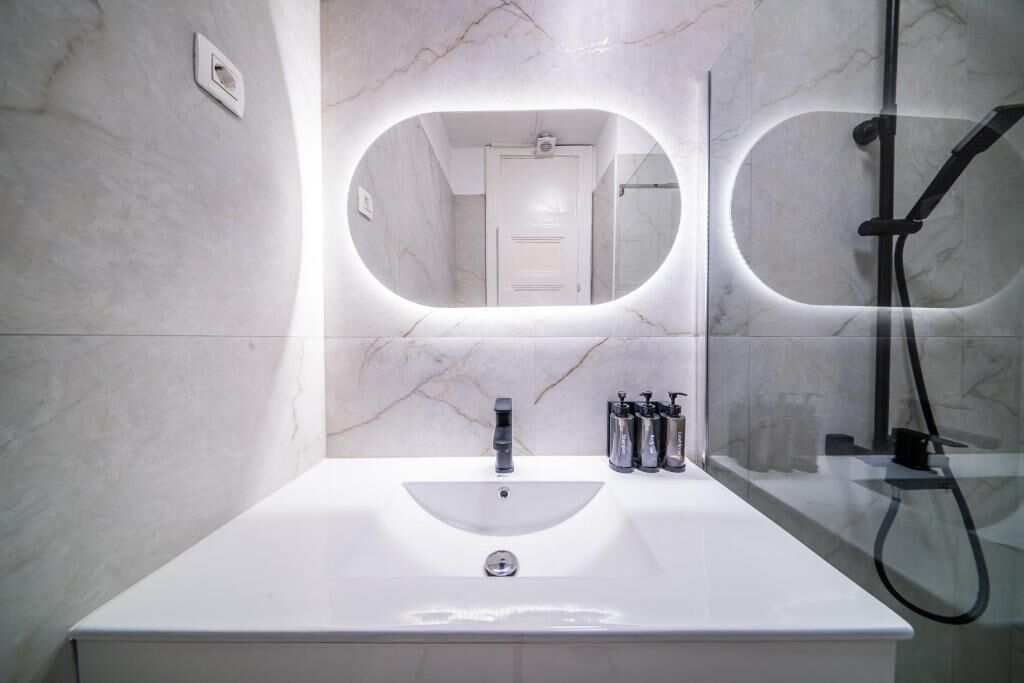 A bathroom with a white sink, a mirror with integrated lighting, a shower, and a door leading to another room.