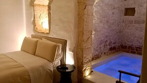 A room with a bed, a small table, and a stone wall with a blue pool. - Masseria Camerlengo (CASTELLANA GROTTE)