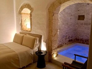 A room with a bed, a small table, and a stone wall with a blue pool. - Masseria Camerlengo (CASTELLANA GROTTE)