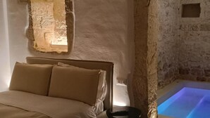 A bedroom with a bed, a nightstand, a stone wall, and a lit blue pool. - Masseria Camerlengo (CASTELLANA GROTTE)