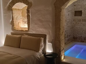 A bedroom with a bed, a nightstand, a stone wall, and a lit blue pool. - Masseria Camerlengo (CASTELLANA GROTTE)