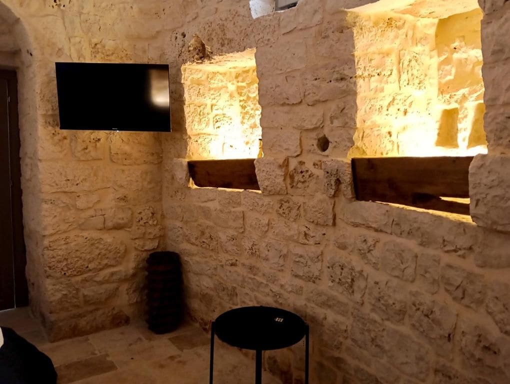 Una stanza con pareti in pietra, una televisione a schermo piatto montata al muro, un tavolo rotondo nero e due finestre con stipiti in legno.