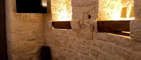 Habitación con paredes de piedra, un televisor de pantalla plana montado en la pared, una mesa redonda negra y dos ventanas con marcos de madera.