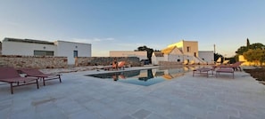 A pool area with lounge chairs, a stone wall, and modern buildings in the background.