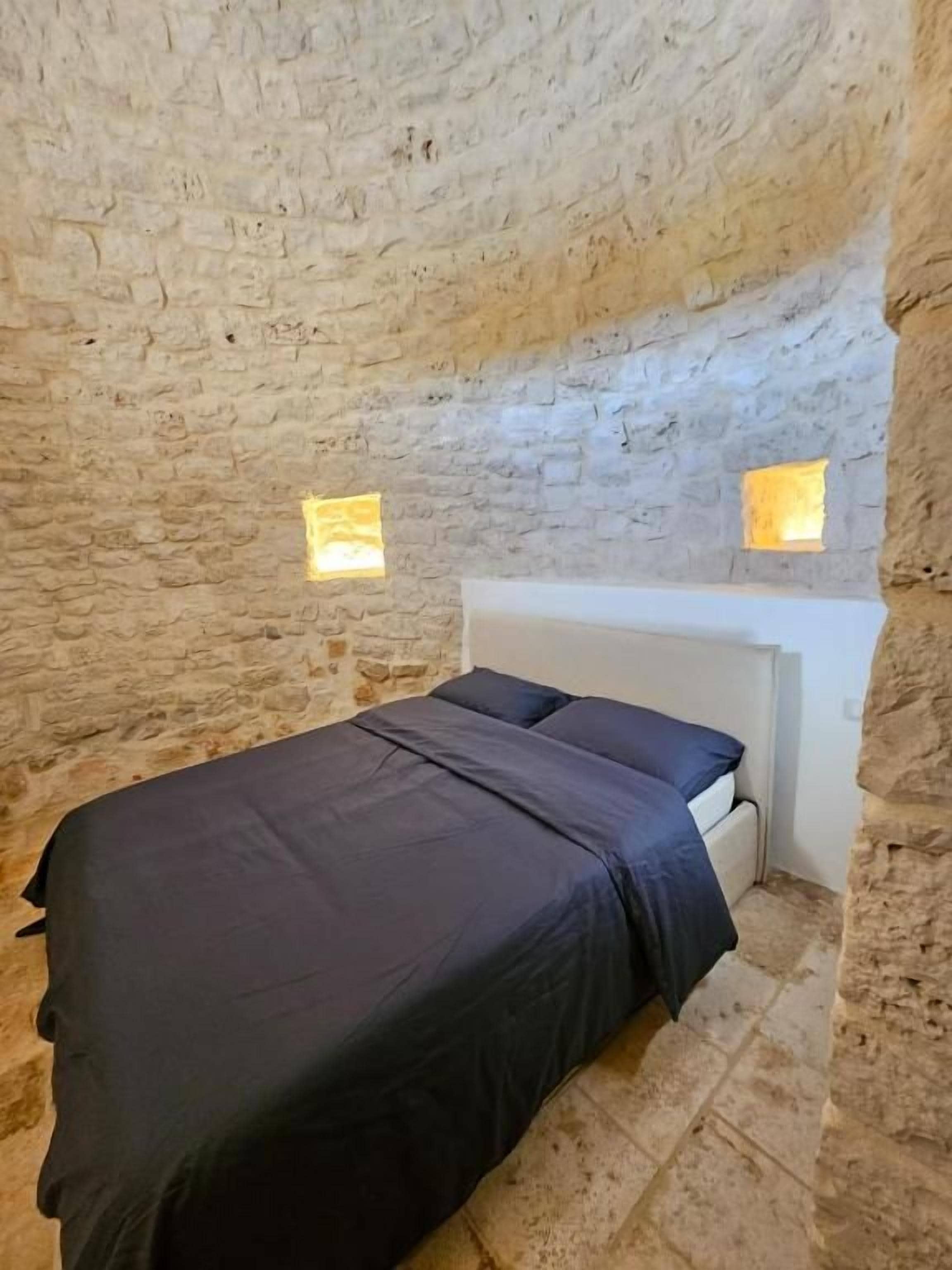Una stanza con parete in pietra, un letto con testiera bianca e due piccole finestre.