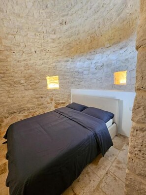 A room with a stone wall, a bed with a white headboard, and two small windows.