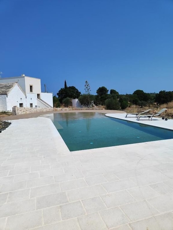 A swimming pool with a white tiled deck, a white building, and trees in the background.
