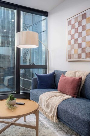 A modern living room with a large window, a blue sofa, a round coffee table, and a floor lamp. - LaneStay - 1BR Southbank Skyline Stay, Pool & Gym (Southbank)