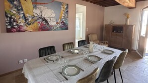 A dining area with a table set for six, wooden beams on the ceiling, and a large floral painting on the wall.
