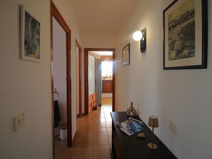 A hallway with a wooden door, a tiled floor, a table with magazines, a lamp, and framed pictures on the walls. - Bright apartment by the beach - 4 guests (Llançà)